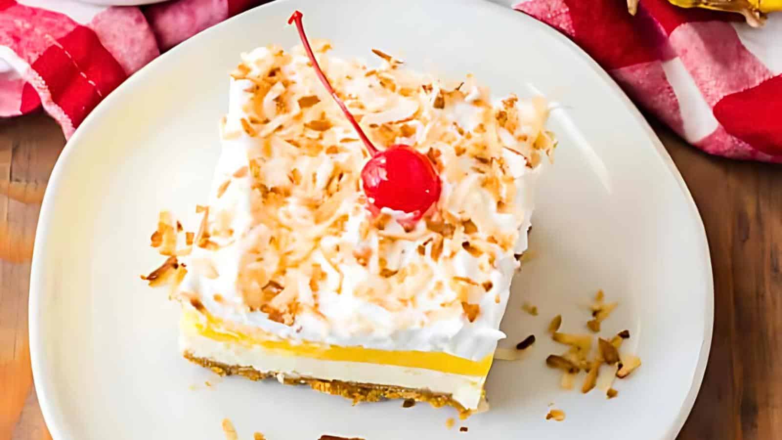 A square slice of layered dessert topped with whipped cream, shredded coconut, and a maraschino cherry on a white plate.