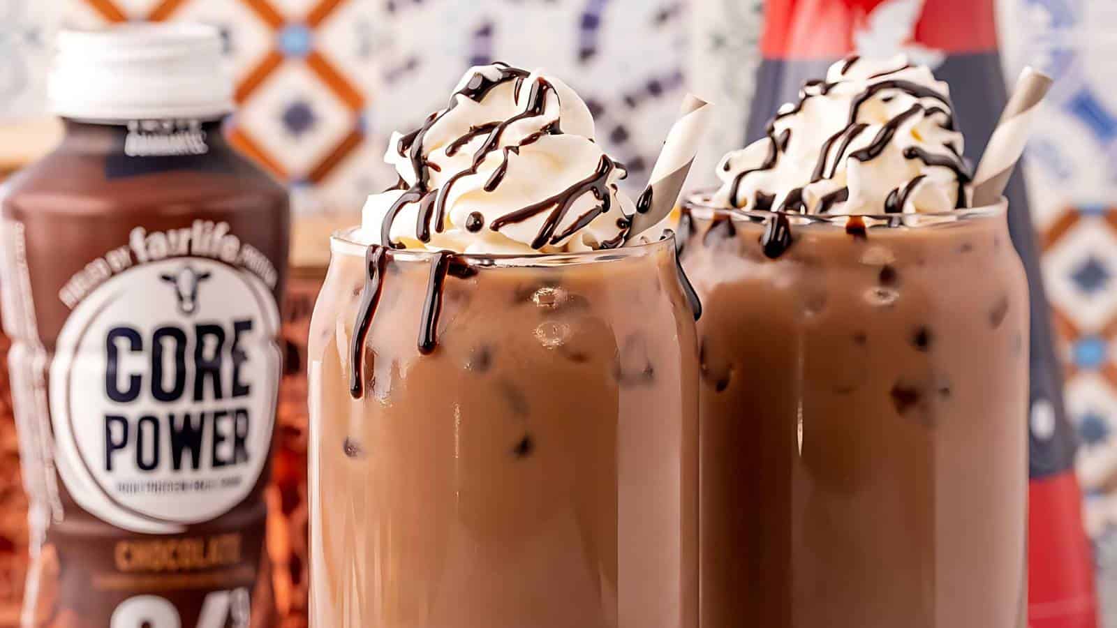 Two iced coffee drinks topped with whipped cream and chocolate drizzle sit next to a bottle of Core Power chocolate protein shake, with a checkered background.