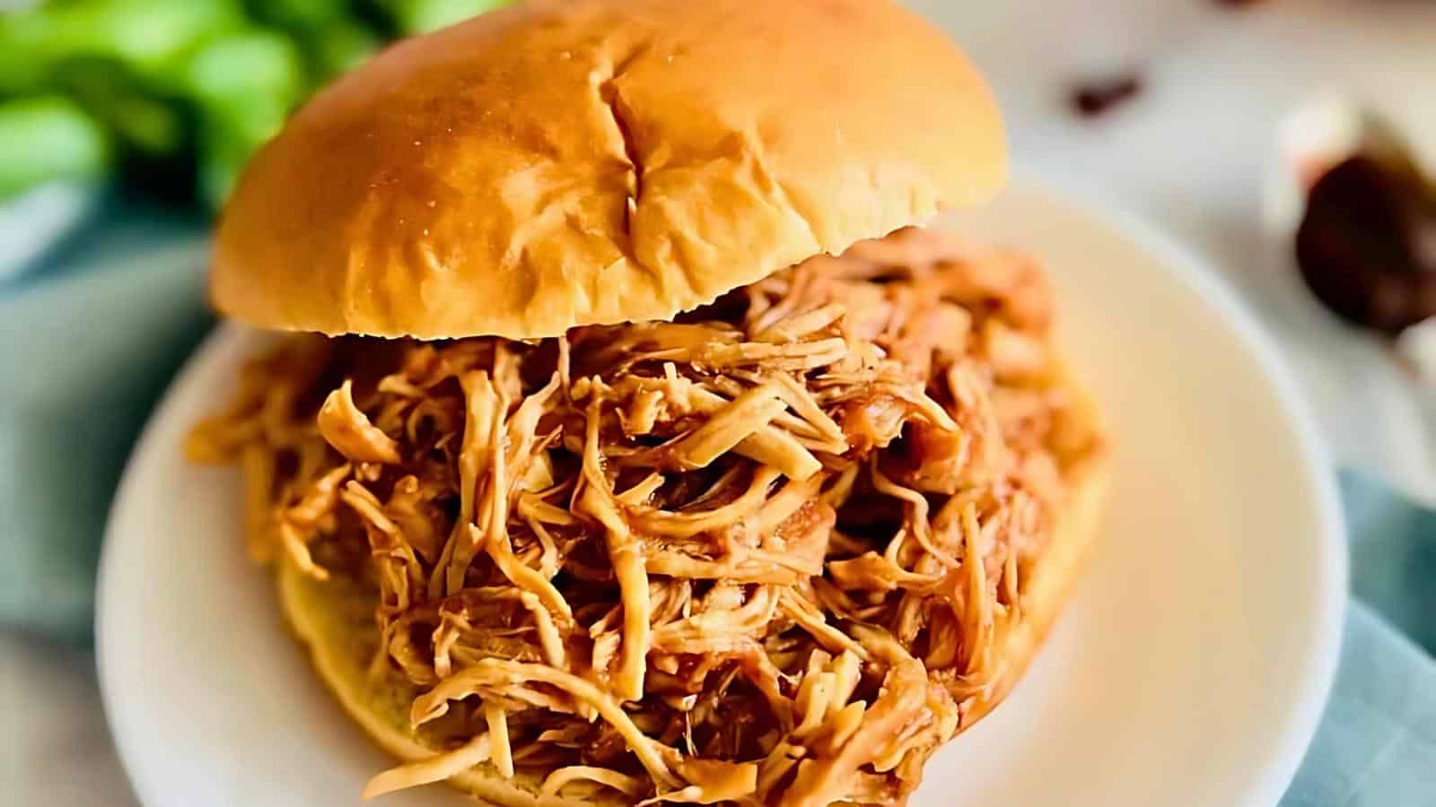A pulled chicken sandwich on a white plate, with shredded pork spilling out from inside a toasted bun.