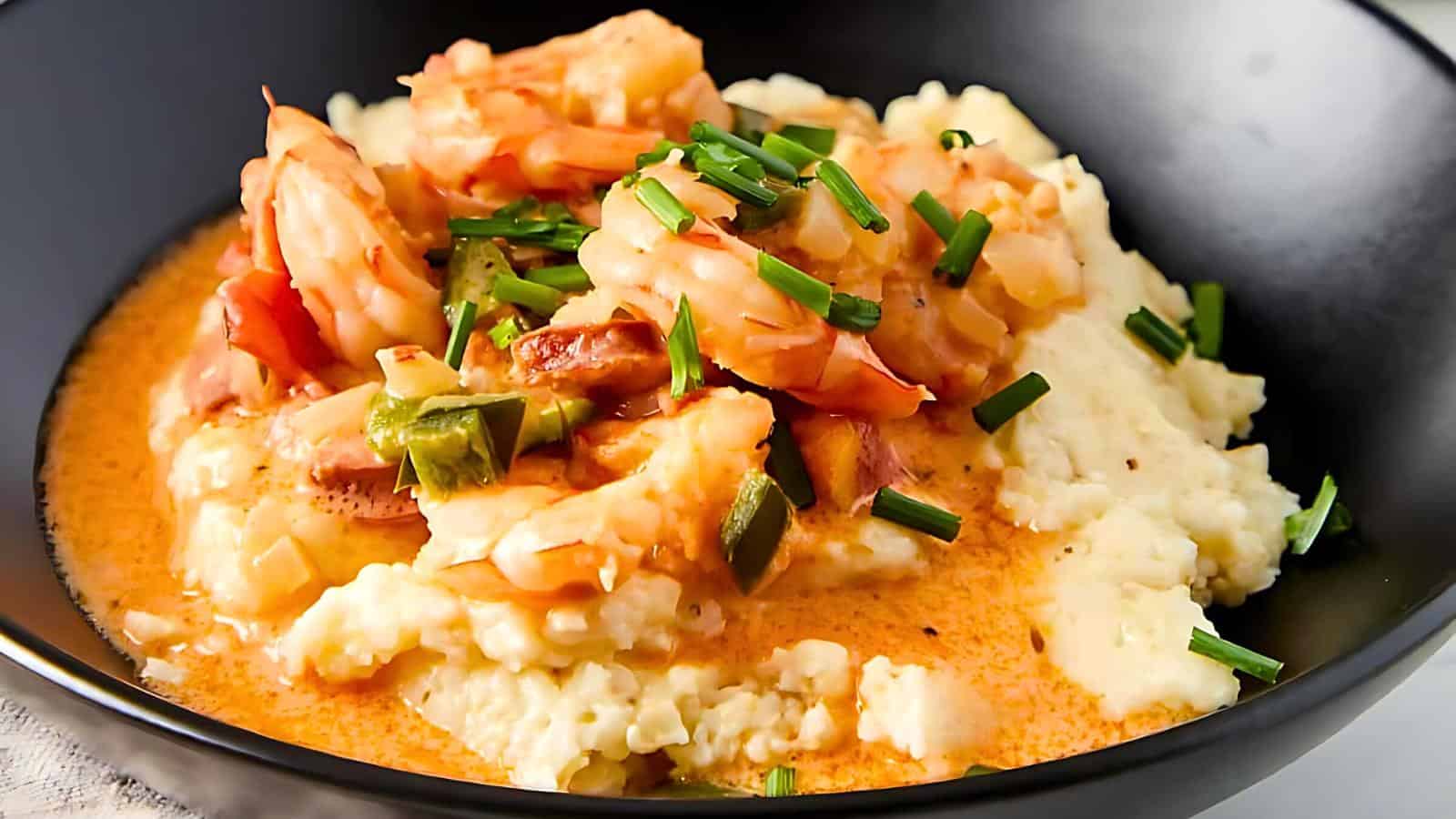 A black bowl with creamy grits, shrimp, diced bell peppers, chives, and a red-tinted sauce.