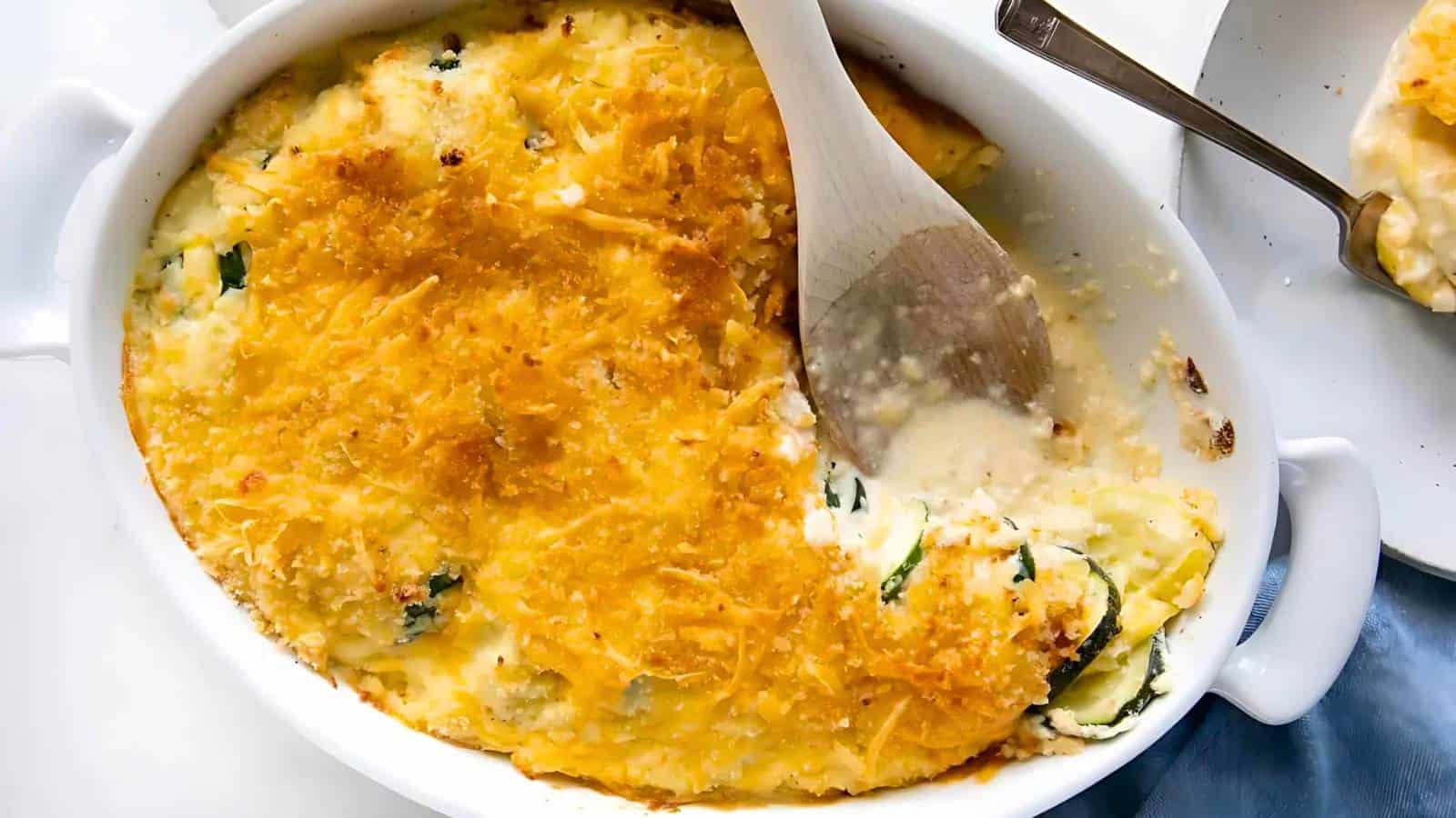 A white oval baking dish filled with a golden-brown casserole, partially served with a wooden spoon, showing a creamy and cheesy interior with visible vegetable layers.