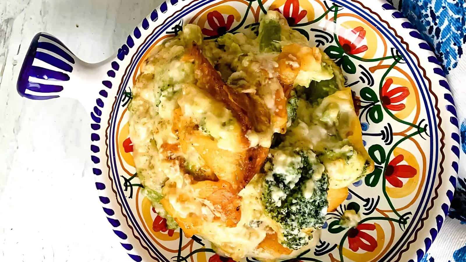 This image shows a colorful plate with a baked dish featuring broccoli florets, gooey melted cheese, and crusty pieces of bread or pasta. The plate has a vibrant floral design, and a blue-patterned cloth is partially visible in the upper right corner.