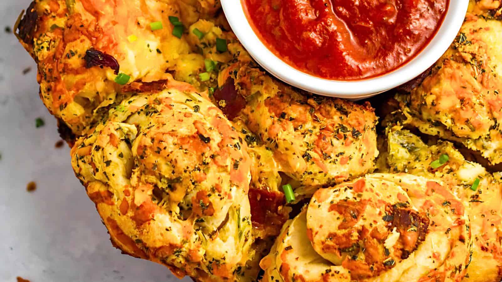 Close-up of savory pull-apart bread rolls sprinkled with herbs and cheese, accompanied by a small bowl of red dipping sauce in the center. The rolls appear golden and fluffy, with bits of green and red ingredients visible.