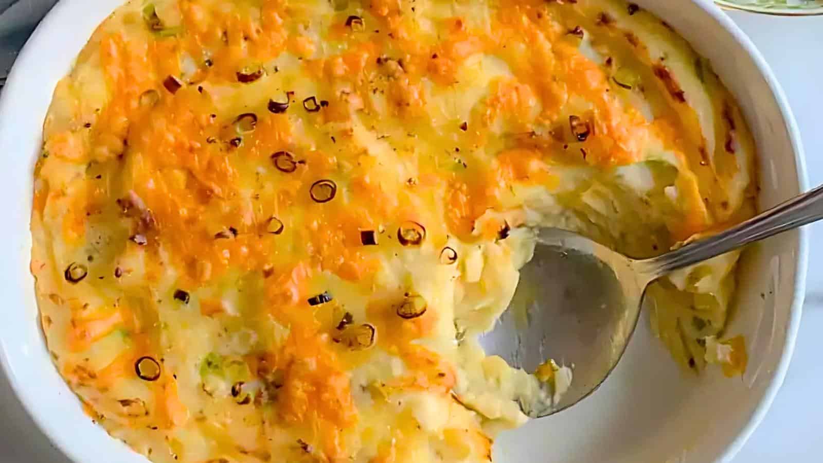 A white bowl filled with a baked dish topped with melted cheese and garnished with sliced spring onions. A spoon is scooping out a portion, revealing a creamy texture underneath.