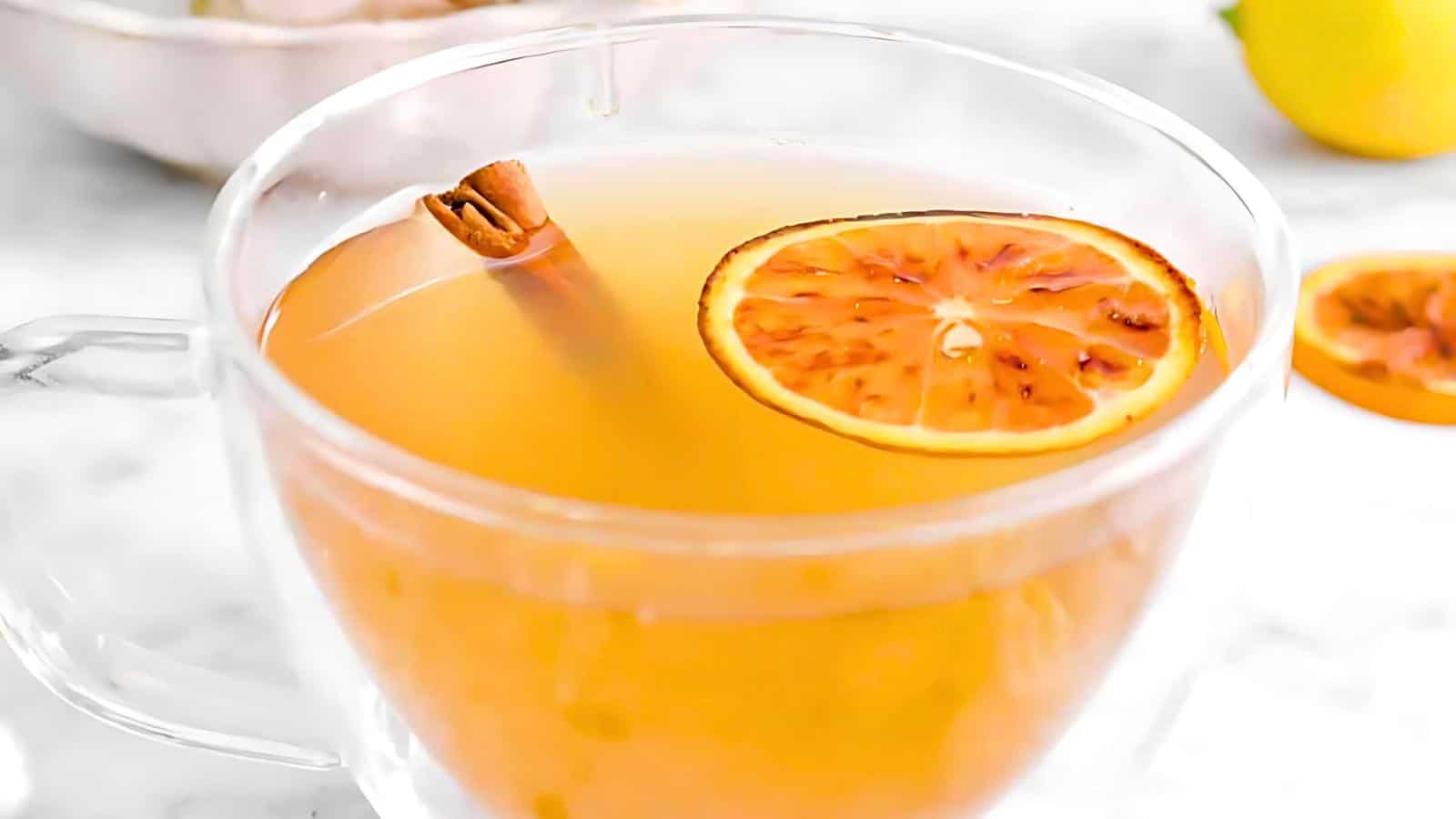 A clear glass mug filled with a light orange beverage, garnished with a slice of dried orange and a cinnamon stick.