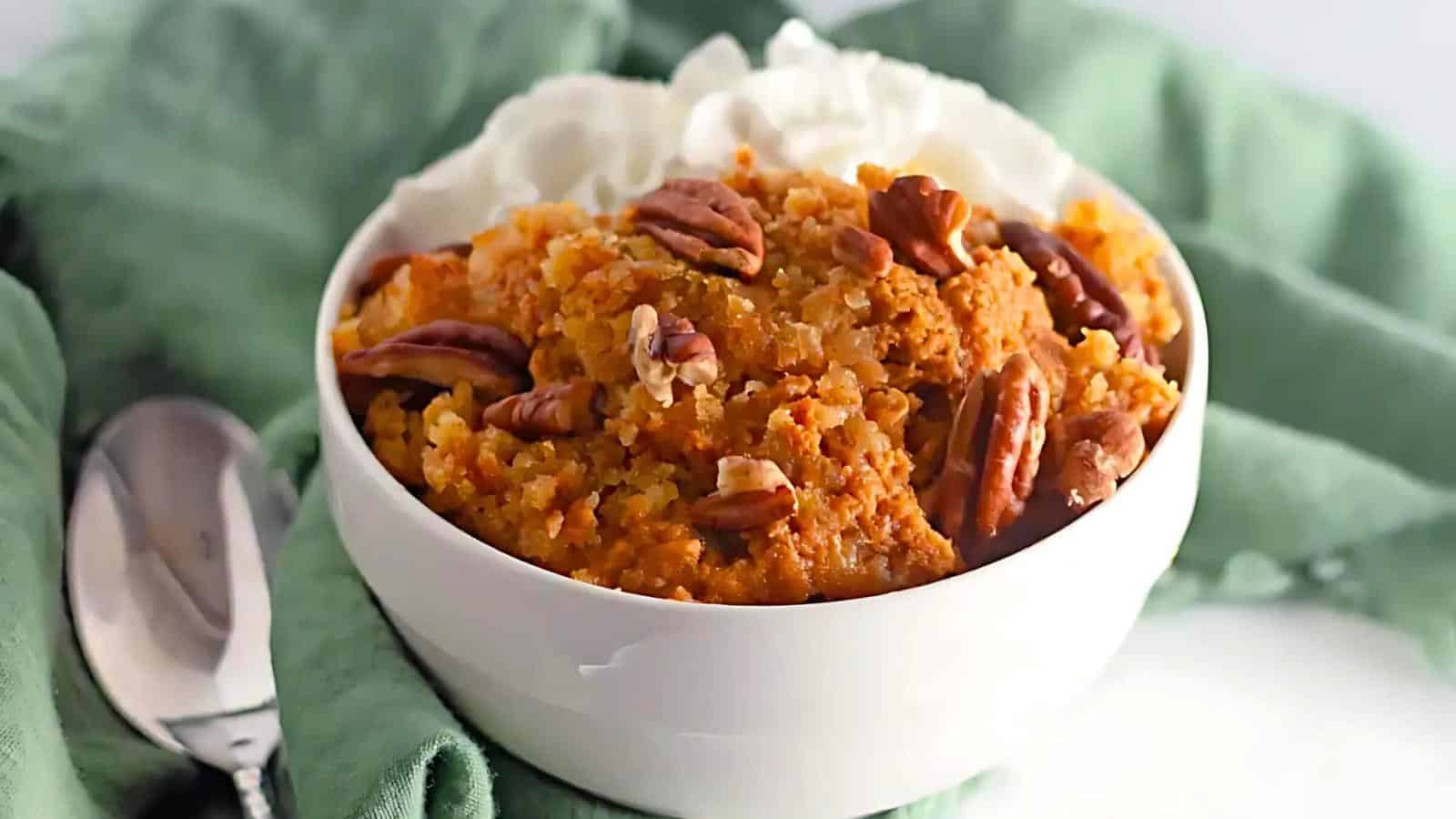 A white bowl filled with sweet potato casserole, topped with pecans and dollops of whipped cream, is placed on a green cloth napkin. A silver spoon lies to the left of the bowl. The setting is bright, with white surfaces in the background.