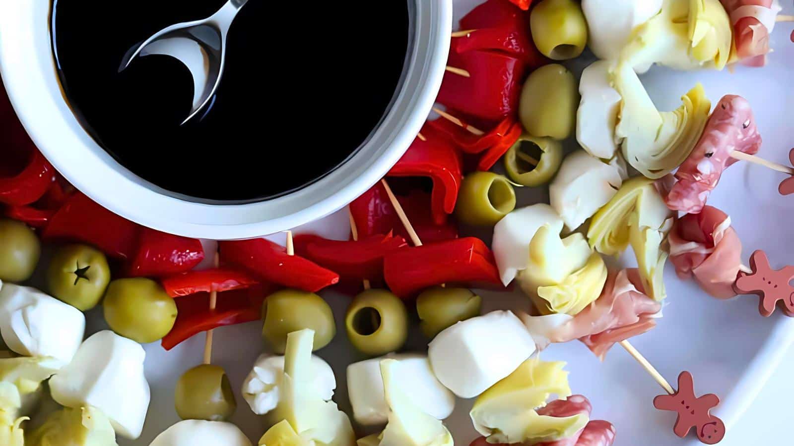 A platter with skewered olives, mozzarella, artichokes, red peppers, and prosciutto. A small bowl of dark dipping sauce with a spoon on the side.