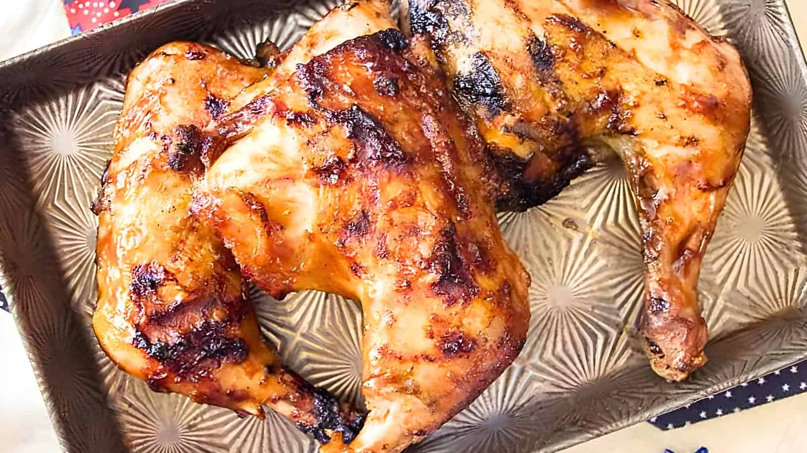 Two grilled chicken halves with a crispy, browned exterior on a textured metal baking tray.