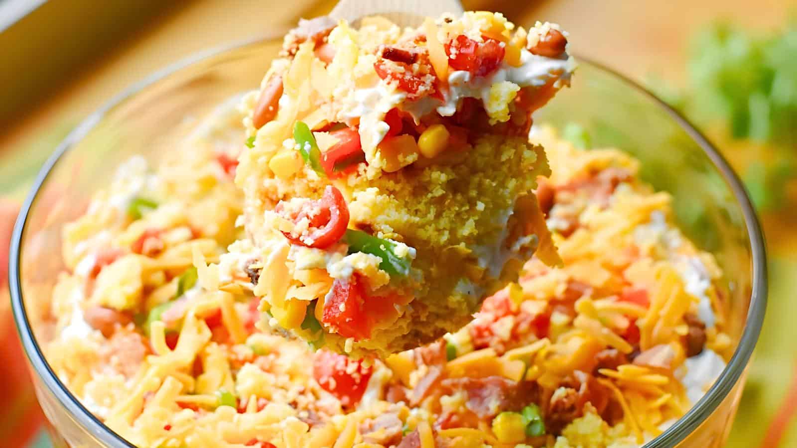 A glass bowl containing layers of cornbread, cheese, tomatoes, corn, green onions, and bacon.