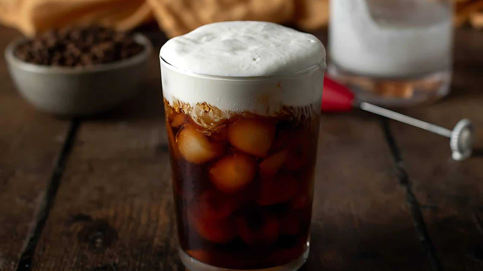 A glass of iced coffee with thick foam on a dark wooden table. A bowl of coffee beans and a frother in the background.