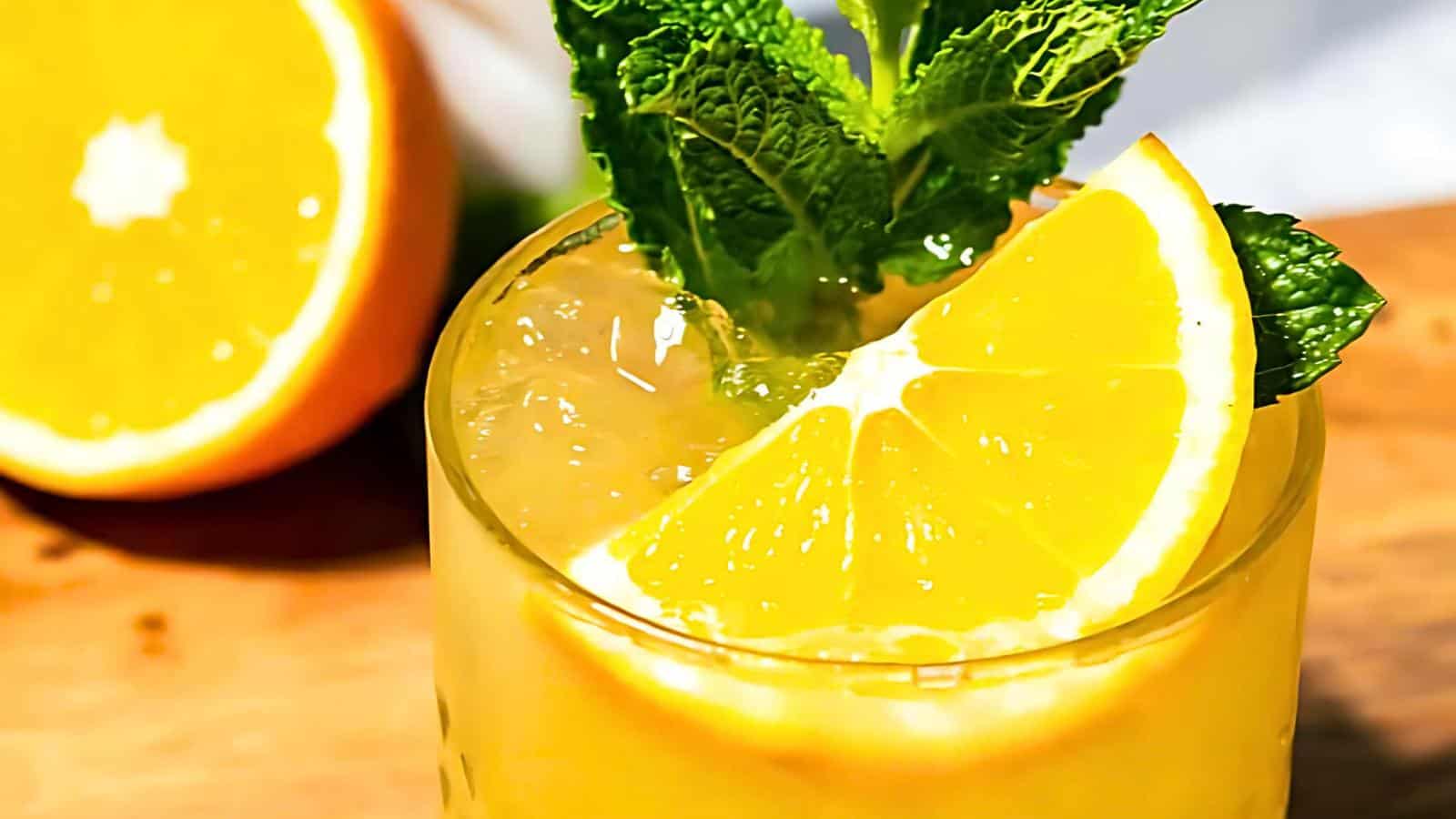 A glass of mocktail with ice, garnished with orange slices and fresh mint leaves, on a wooden surface, with a halved orange in the background.