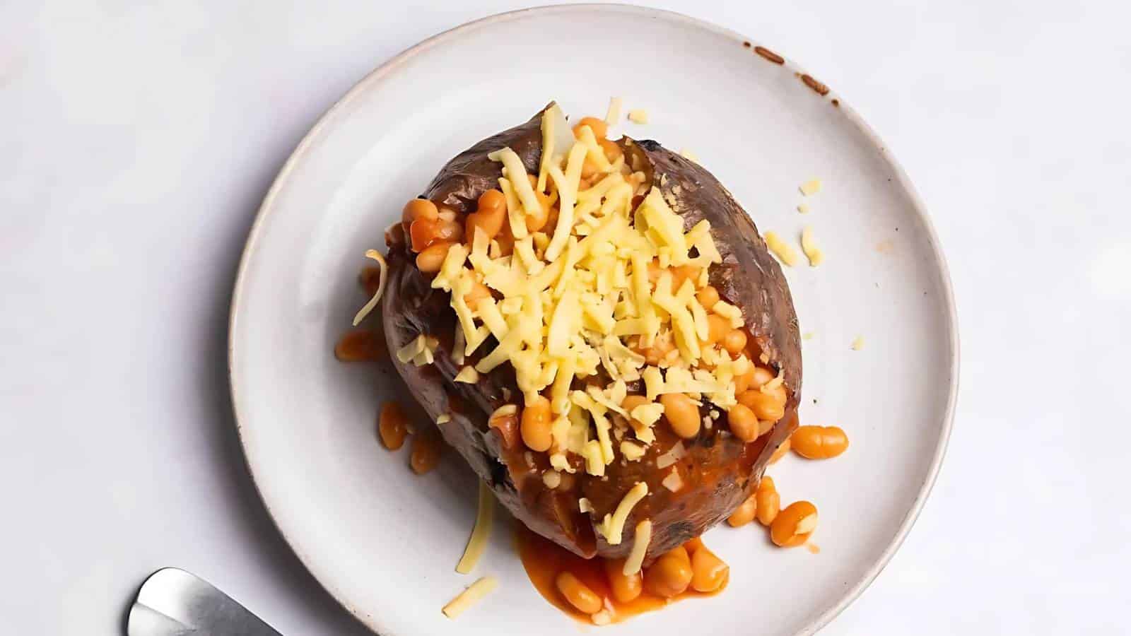 A baked potato topped with baked beans and shredded cheese on a white plate, with a spoon partially visible beside it.