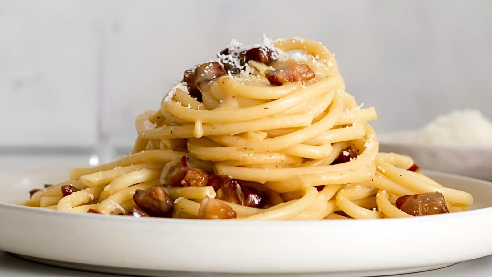 A plate of spaghetti carbonara is topped with grated cheese and pieces of crispy pancetta, with a creamy sauce coating the pasta. The dish is set against a simple, light background, focusing on the food's rich and inviting textures.