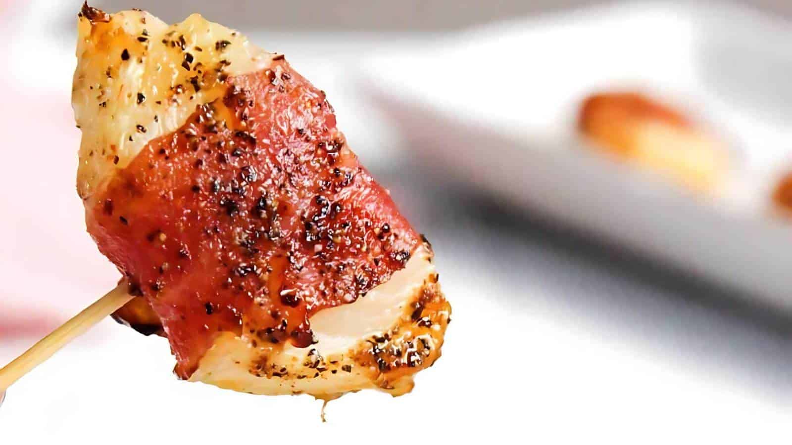 A bacon-wrapped chicken skewer seasoned with pepper, set against a blurred white rectangular plate.