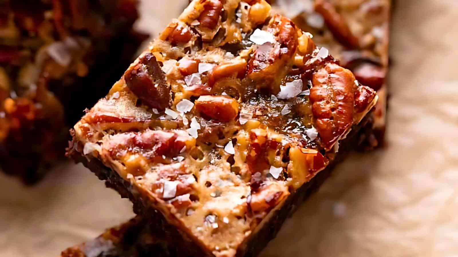 Close-up of a pecan bar topped with flaky sea salt. The bar appears rich and chewy, featuring a mix of dark and caramelized colors from the nuts and syrup. A second bar is partially visible in the background, all placed on a parchment-lined surface.
