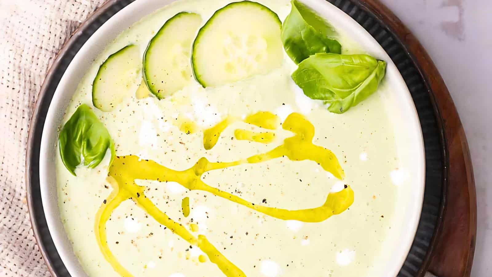 A bowl of green soup garnished with slices of cucumber, basil leaves, a drizzle of yellow oil, and a sprinkle of seasoning.