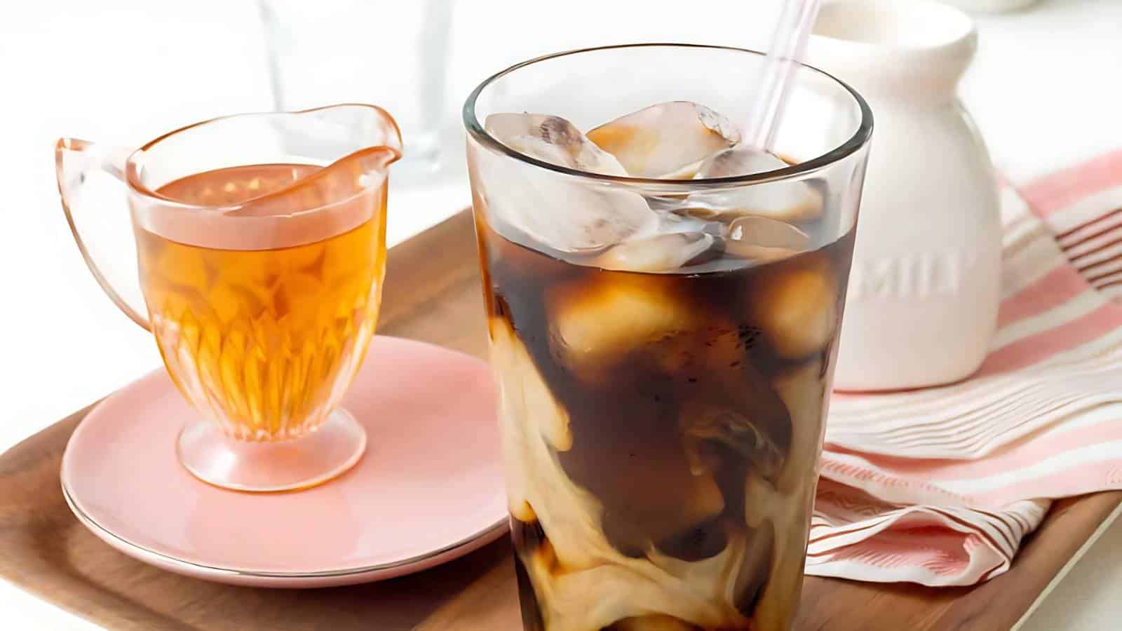 A glass of iced coffee with milk swirling inside sits on a wooden tray next to a small jug of amber-colored liquid and a white milk container. A pink striped cloth is also placed on the tray.