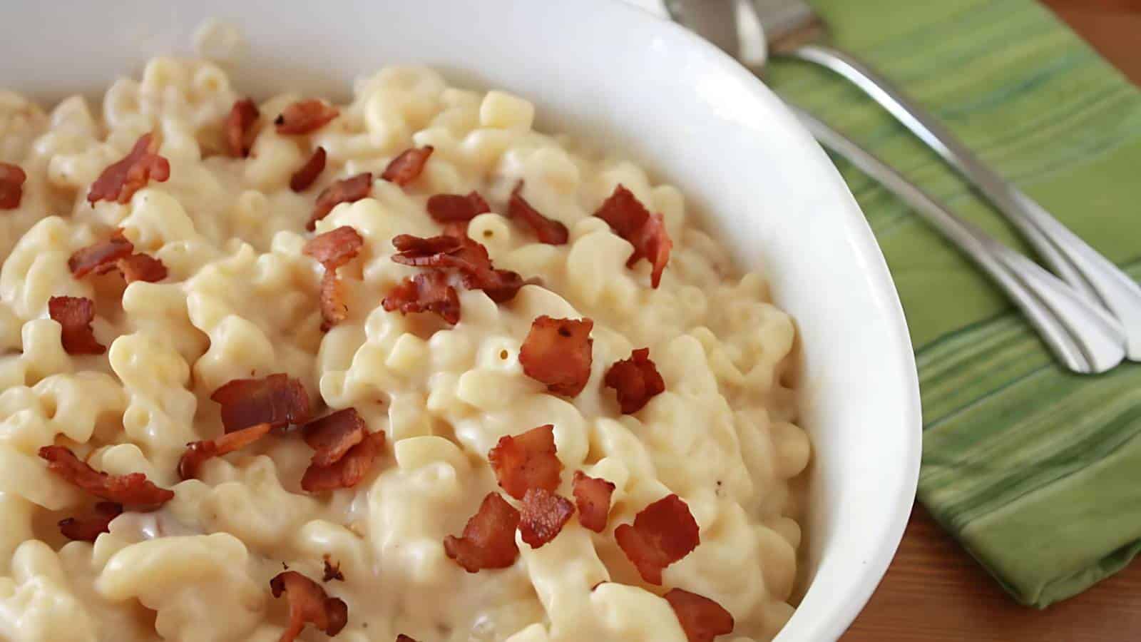 A white bowl filled with creamy macaroni and cheese topped with crispy bacon bits. In the background, there is a green striped napkin and two silver spoons.