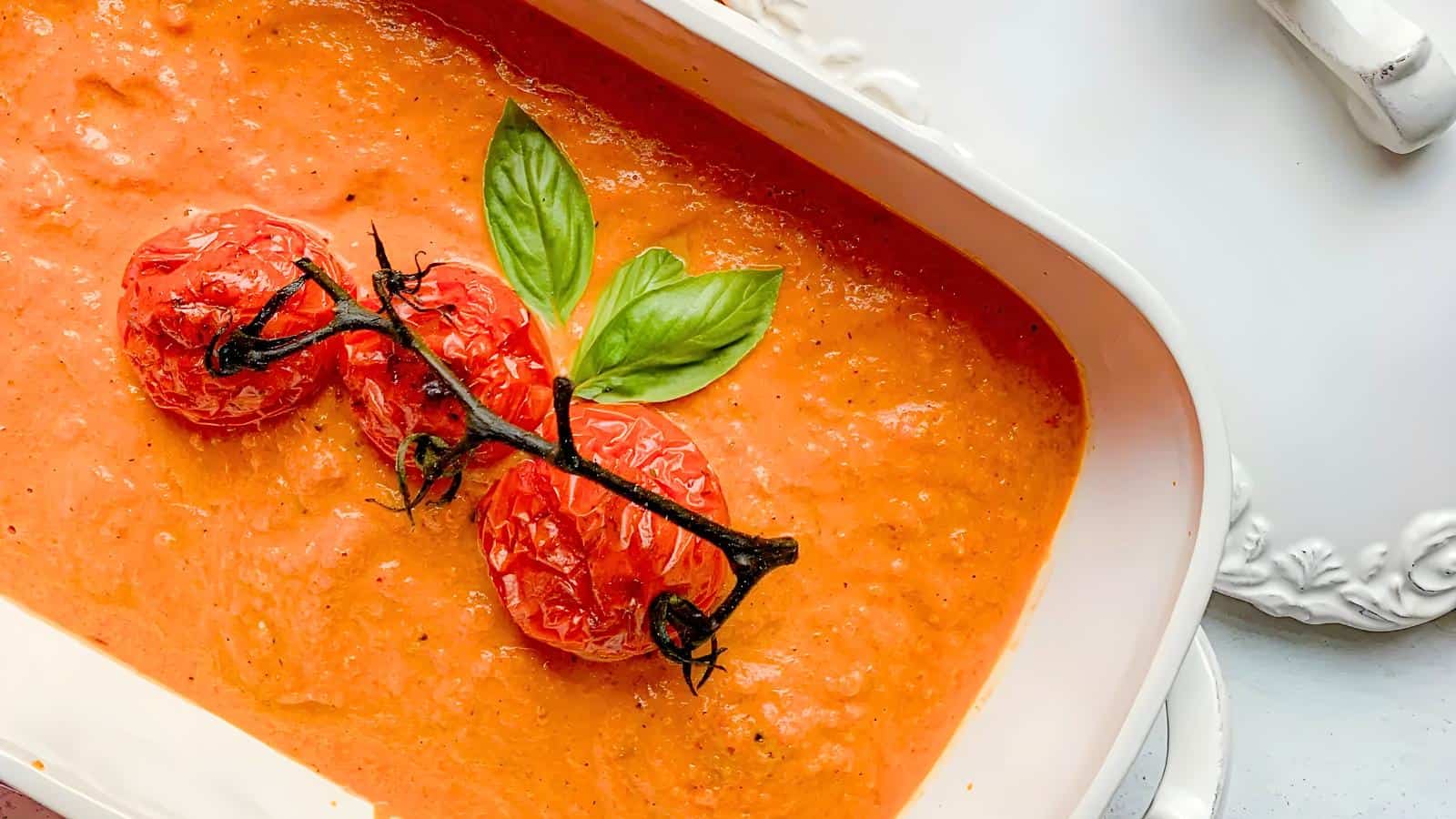 A dish of tomato soup, garnished with three roasted tomatoes on the vine and fresh green basil leaves.