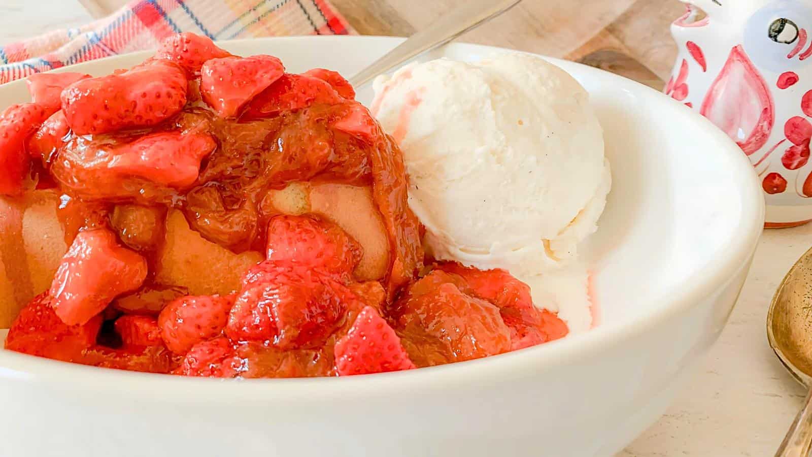 A dessert bowl featuring a baked treat topped with glazed strawberries and a scoop of vanilla ice cream. The dish is set on a table with a plaid cloth and an owl-shaped container in the background.