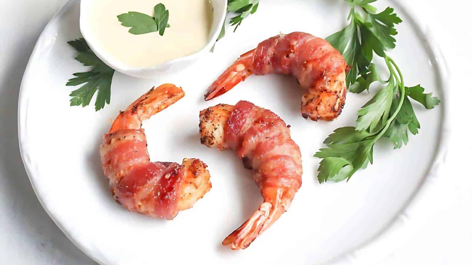 Three bacon-wrapped shrimp, sprigs of parsley, and a small dish of creamy sauce on a white plate.