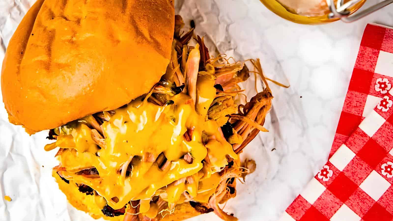 A sandwich with shredded meat and melted cheese on a bun, placed on a white crumpled paper next to a red and white checkered napkin.