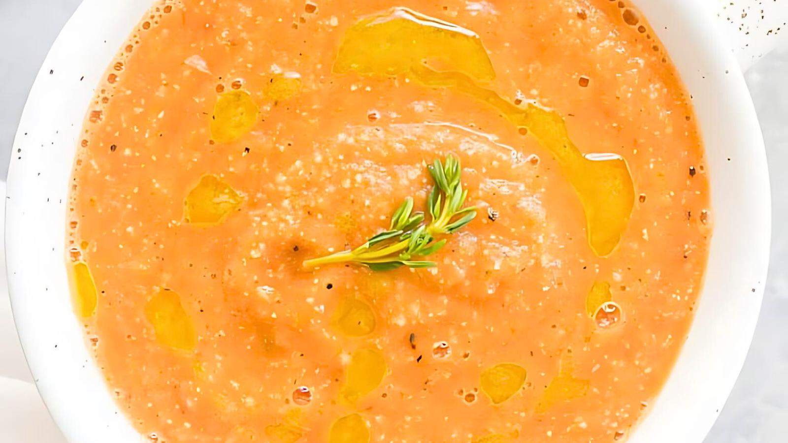 A bowl of creamy tomato soup, garnished with a sprig of fresh thyme and drizzled with olive oil.