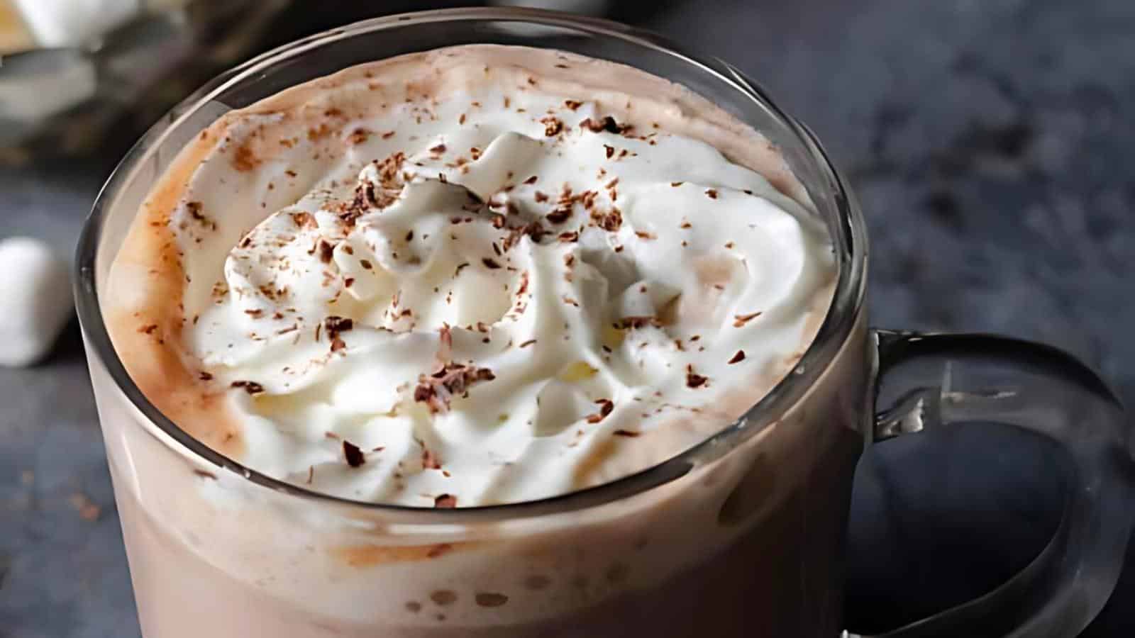 A clear glass mug filled with hot chocolate topped with whipped cream and chocolate shavings.