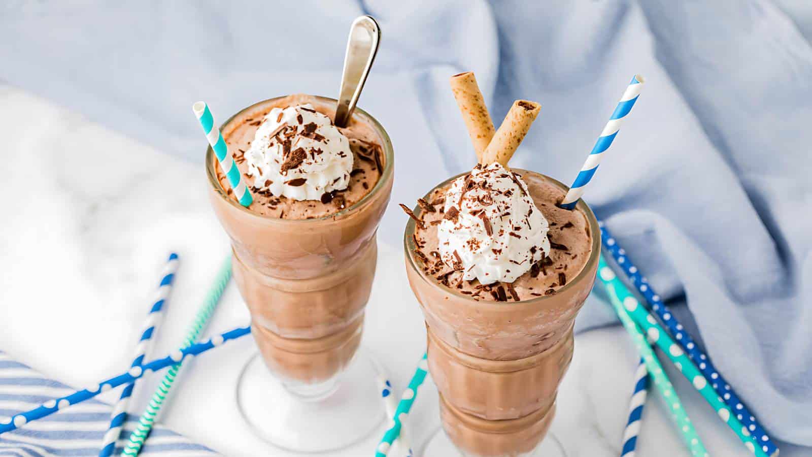 Two tall glasses of chocolate milkshakes topped with whipped cream and chocolate shavings. One has a spoon and a blue-striped straw, while the other has two wafer sticks and a blue-striped straw.