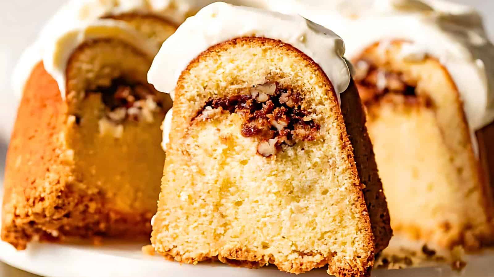 Close-up of a sliced bundt cake with a rich, golden exterior, featuring a visible layer of chopped nuts and a moist filling. The cake is topped with a generous dollop of creamy white icing, and the background showcases additional slices of the same cake.