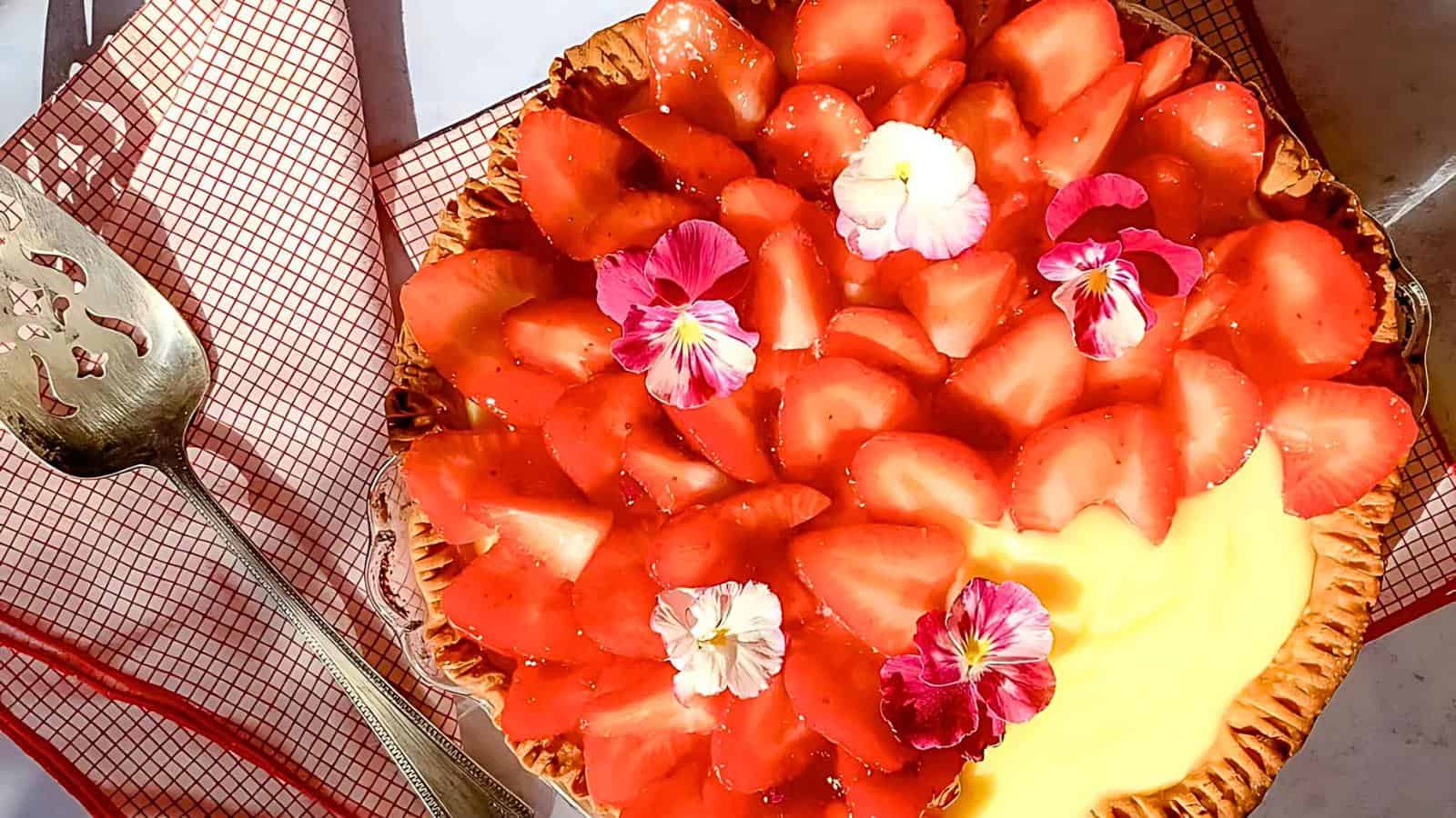A pie topped with fresh strawberries and edible flowers sits next to a pie server on a red and white checkered cloth.