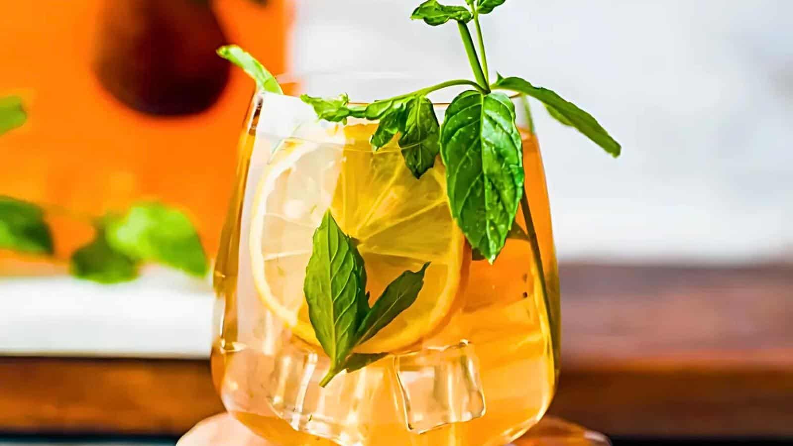 A glass filled with iced tea garnished with a slice of lemon and fresh mint leaves. In the background, an orange-colored beverage and more mint leaves are visible.