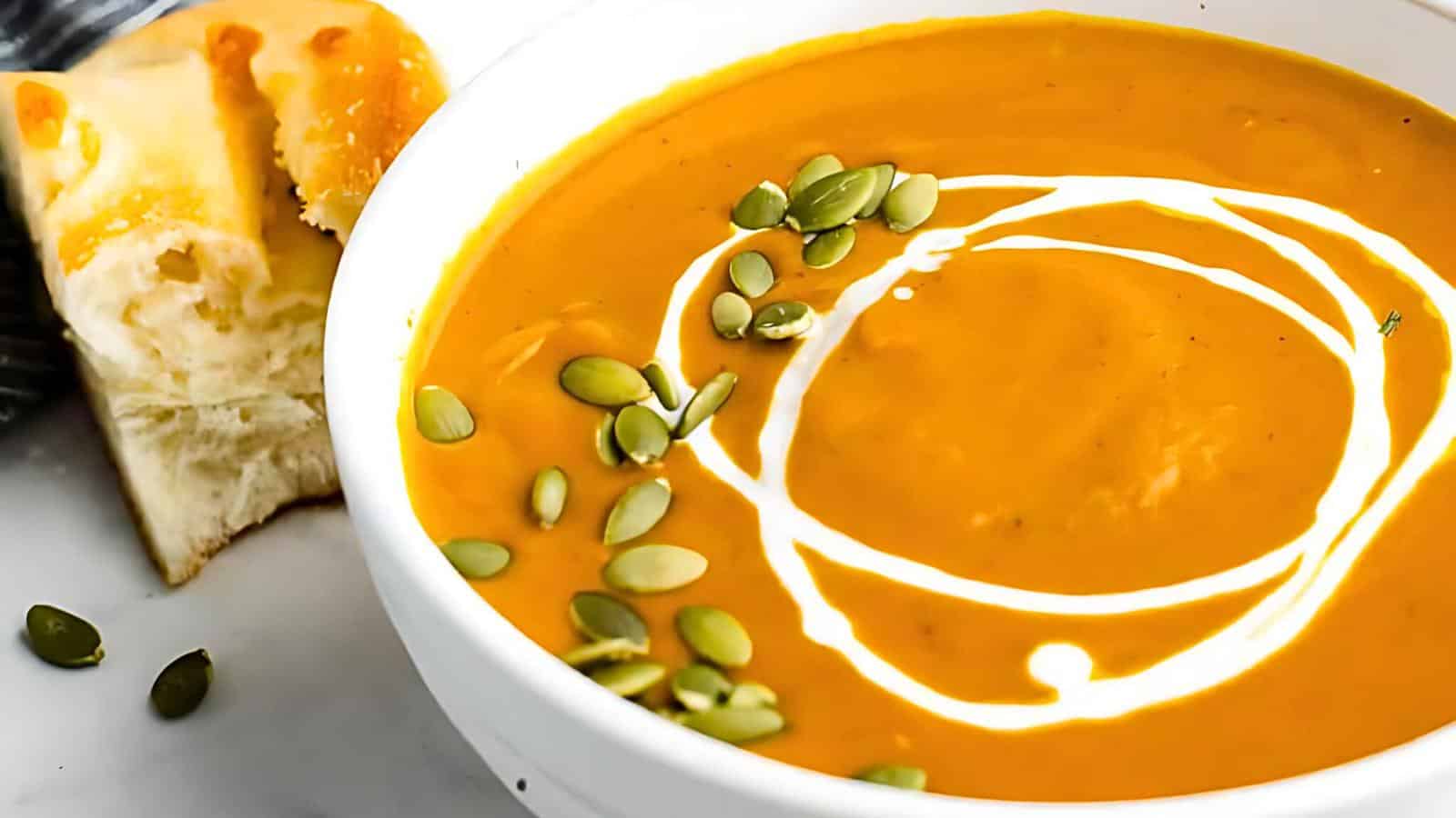 A bowl of creamy orange soup with a swirl of cream and pumpkin seeds, alongside a piece of bread.