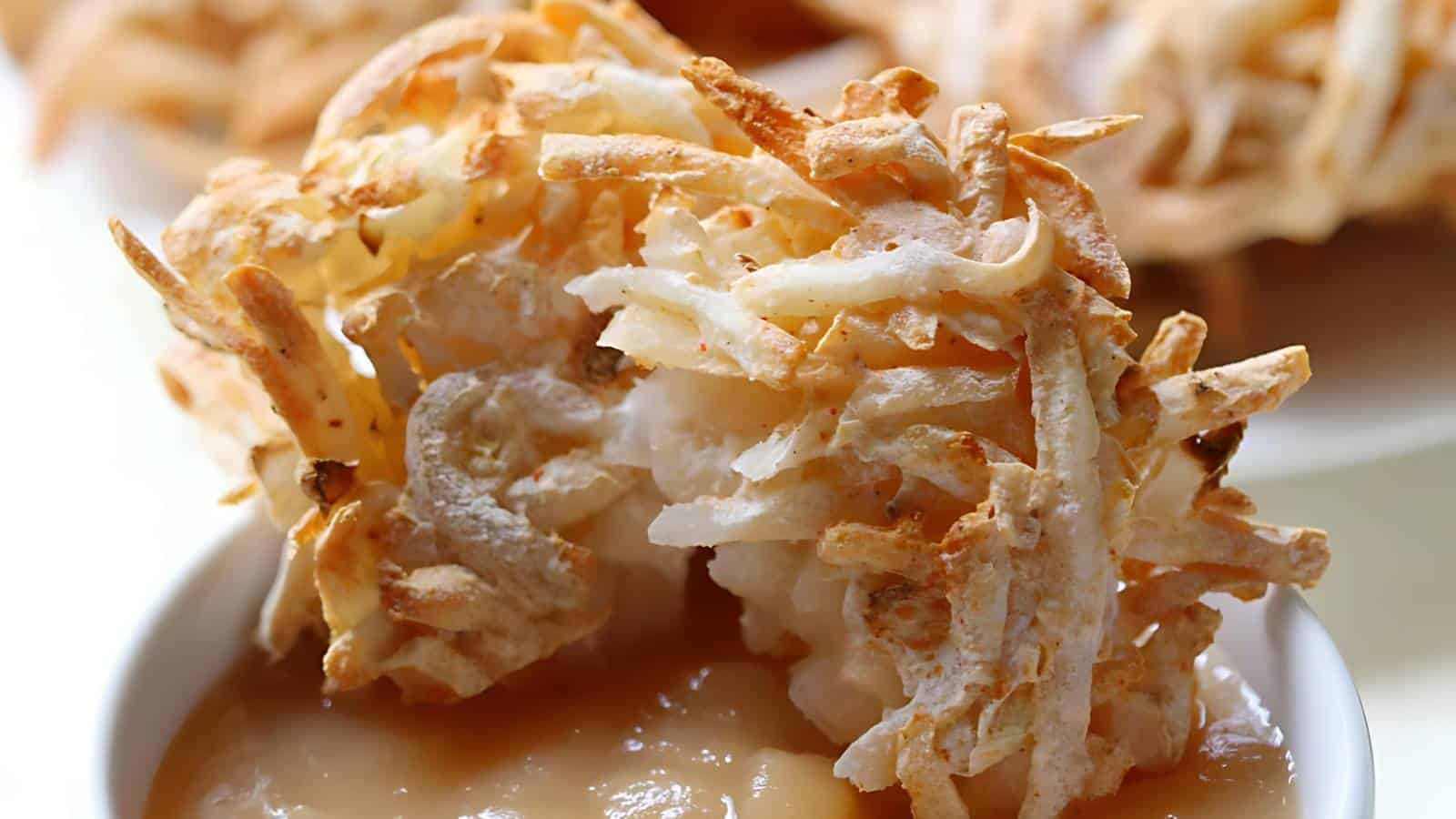 A crispy, golden-brown potato latke with a shredded texture, partially dipped in a light brown sauce within a white dish.