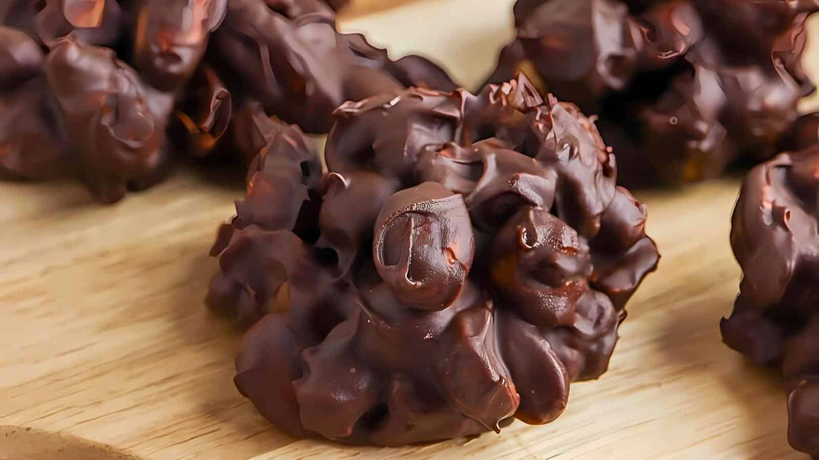 Close-up of clusters of chocolate-covered nuts placed on a wooden surface. The clusters have a rough, textured appearance with shiny, melted chocolate coating the nuts.