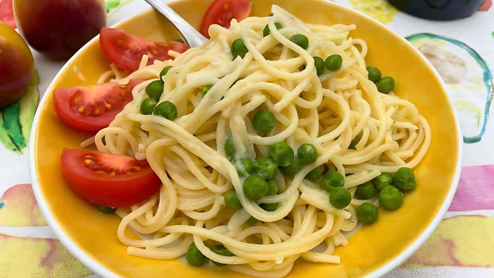 A yellow bowl of spaghetti mixed with green peas, garnished with halved cherry tomatoes, with a fork resting on the side.
