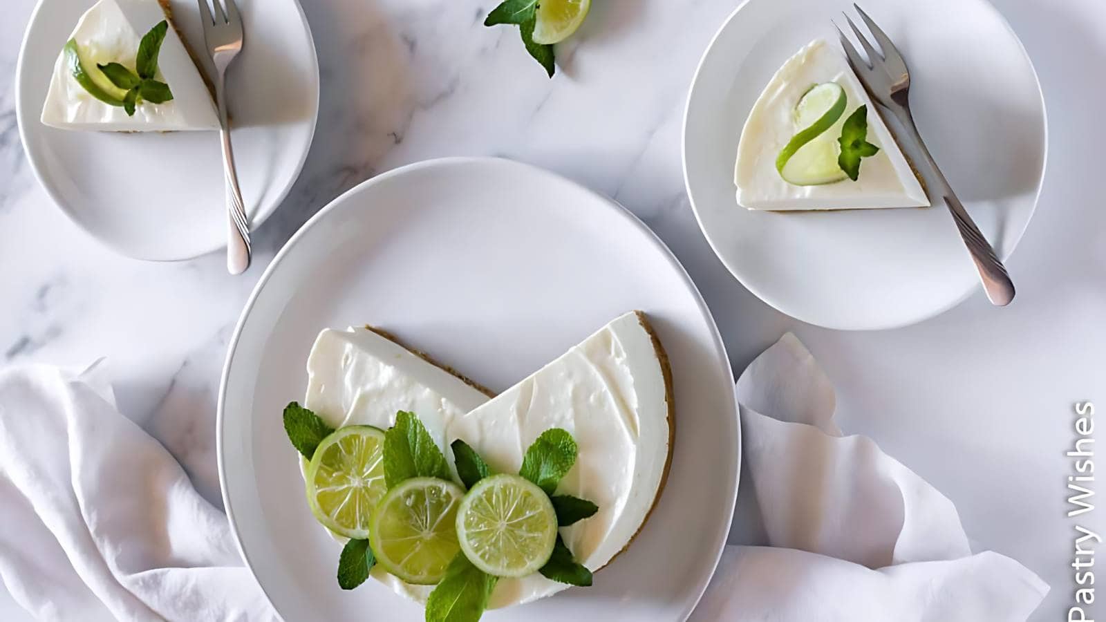 A lime cheesecake with three slices and garnished with lime slices and mint leaves, placed on a white tablecloth.