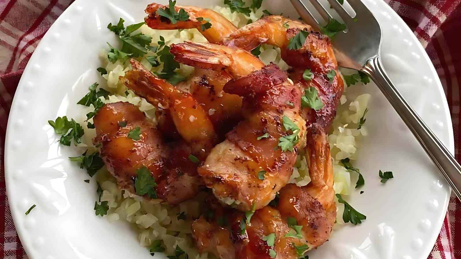 A white plate with bacon-wrapped shrimp on a bed of rice, garnished with chopped parsley. A fork rests on the edge of the plate.
