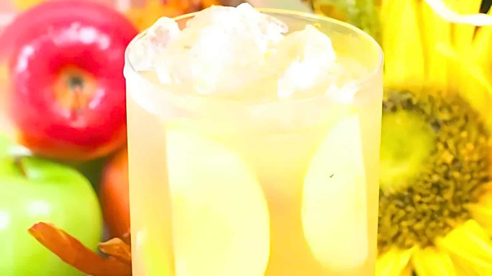 A glass of yellow cocktail with ice cubes, a red apple, a green apple, and a sunflower in the background.