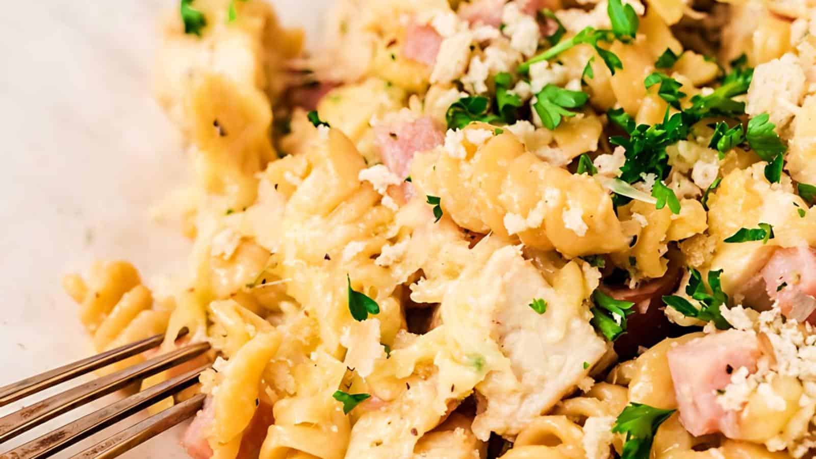 A close-up of a creamy pasta dish with rotini noodles, chunks of ham, and broccoli florets. The pasta is garnished with chopped parsley and grated cheese, creating a colorful and appetizing meal. A fork is partially visible on the left side of the image.