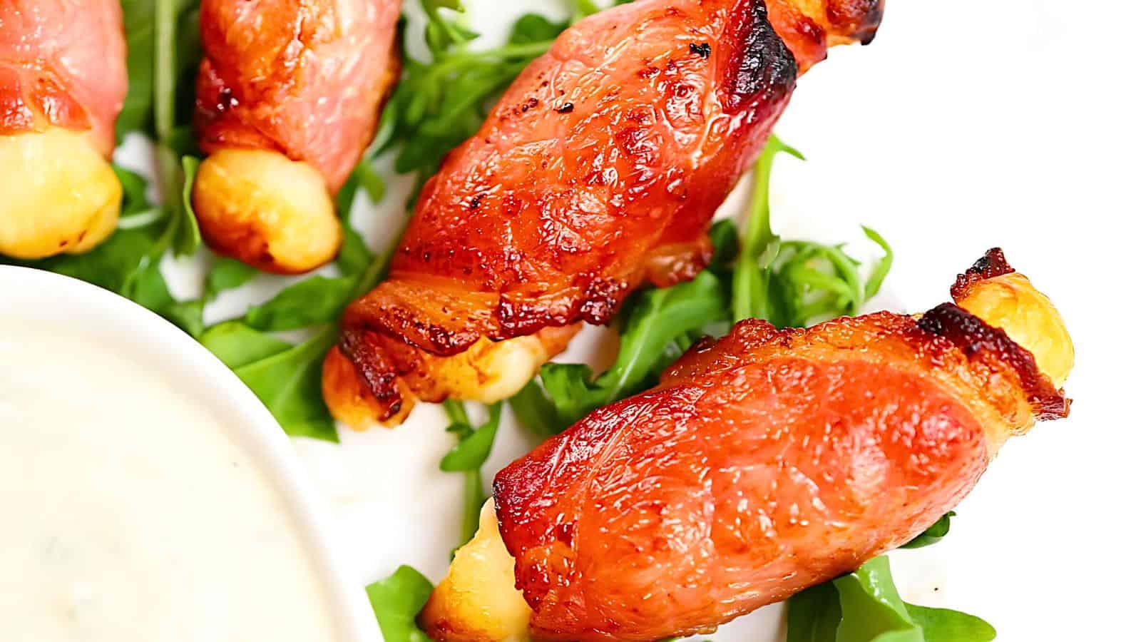 Bacon-wrapped cheese sticks on a bed of fresh green arugula with a side of creamy dipping sauce.
