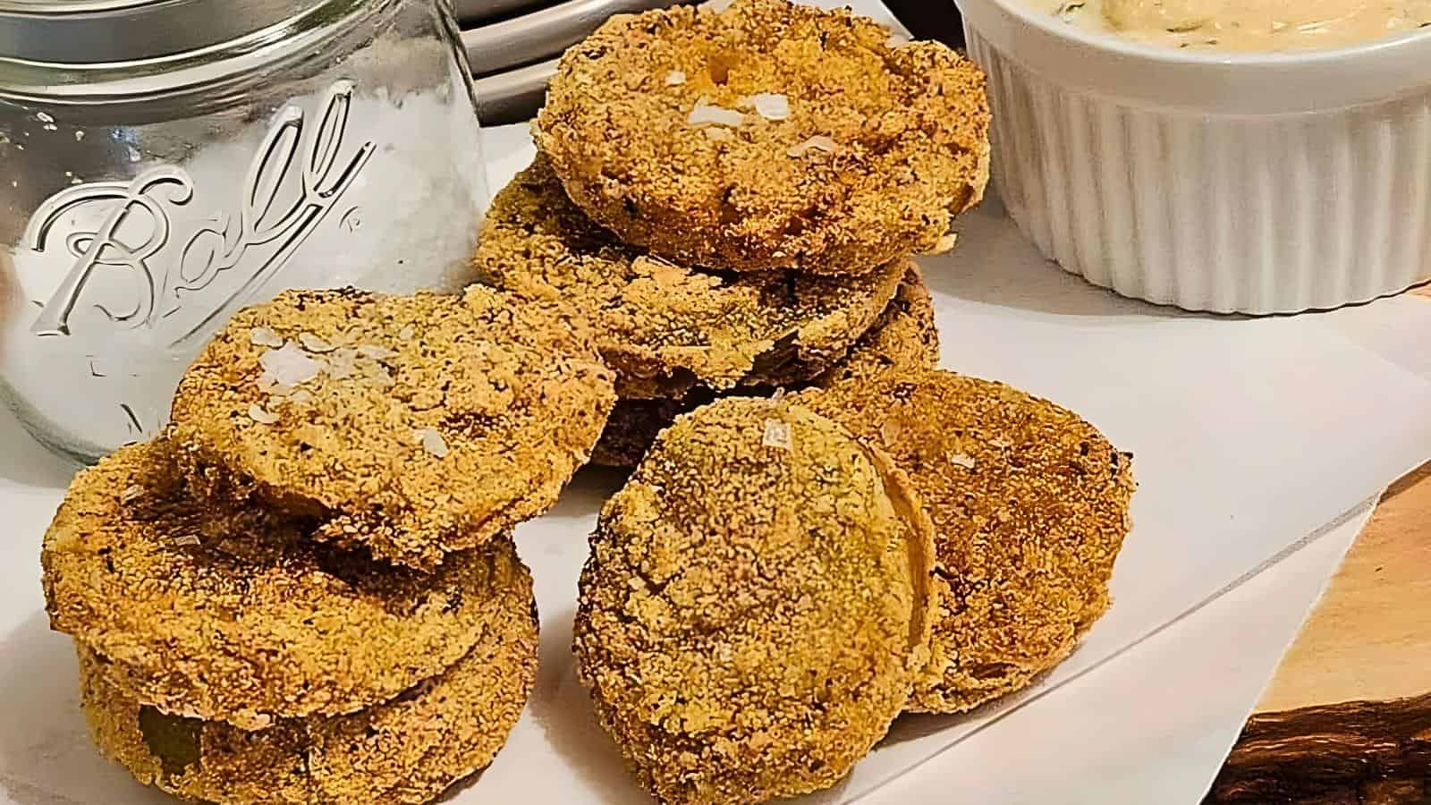 A stack of breaded and fried green tomatoes, a glass jar, and a white ramekin filled with creamy dipping sauce.