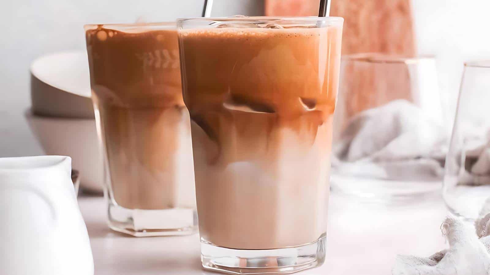 Two glasses of iced coffee on a white surface, surrounded by bowls, a jug, and a white cloth.