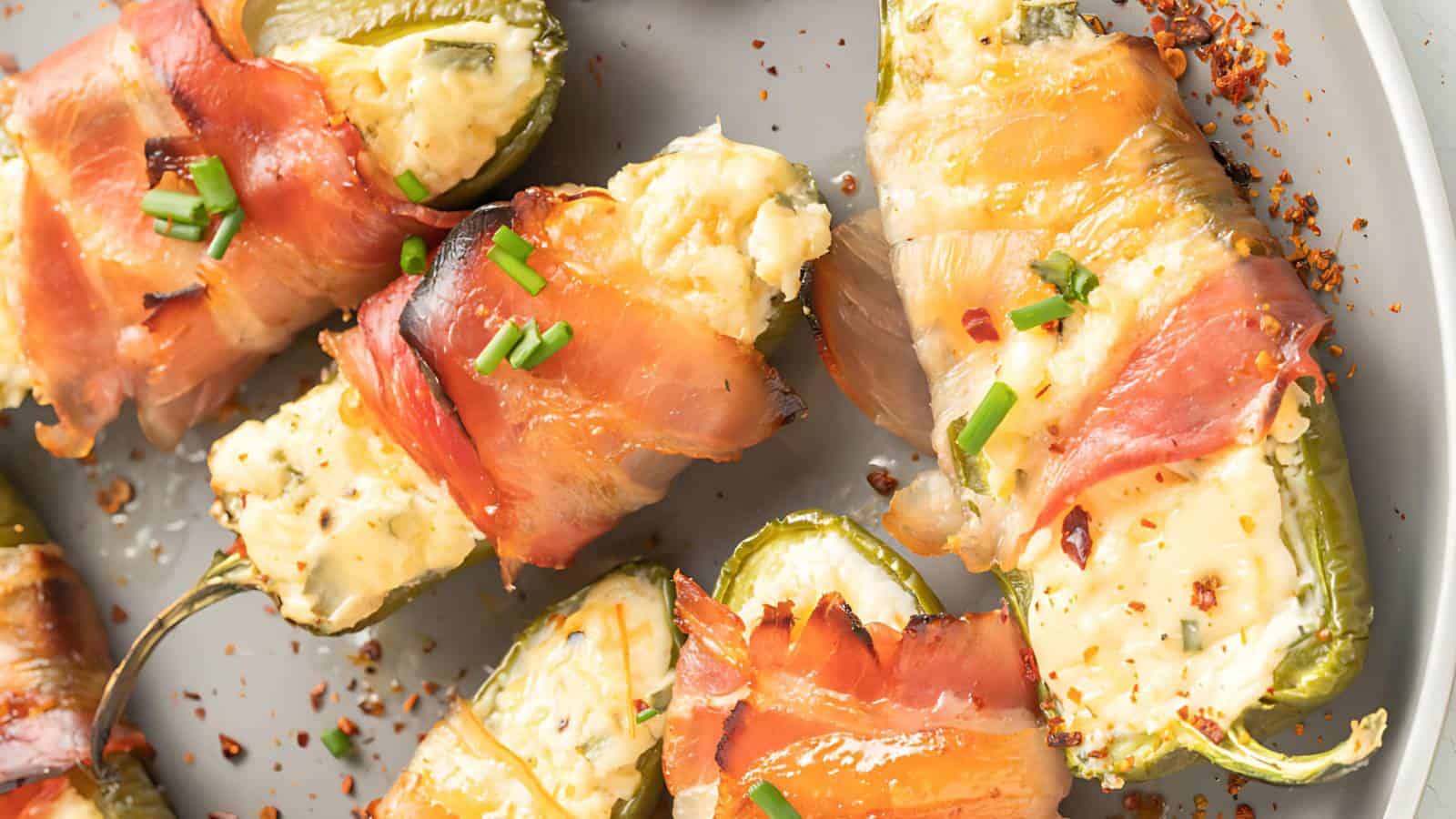 A plate of bacon-wrapped jalapeño poppers filled with cream cheese, garnished with chopped chives and red pepper flakes.
