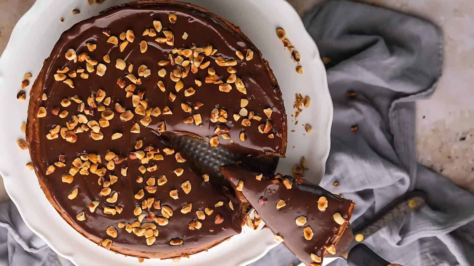 A chocolate cake topped with chopped nuts on a white cake plate, surrounded by gray fabric.