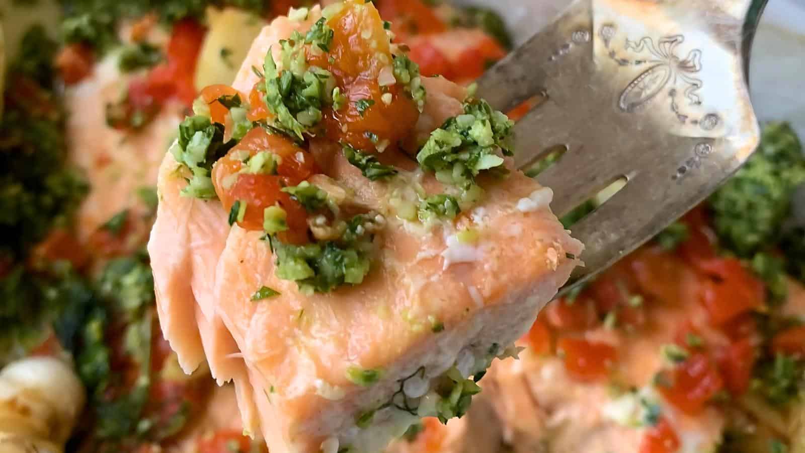 A fork holding a piece of cooked salmon topped with green herbs and diced red tomatoes.