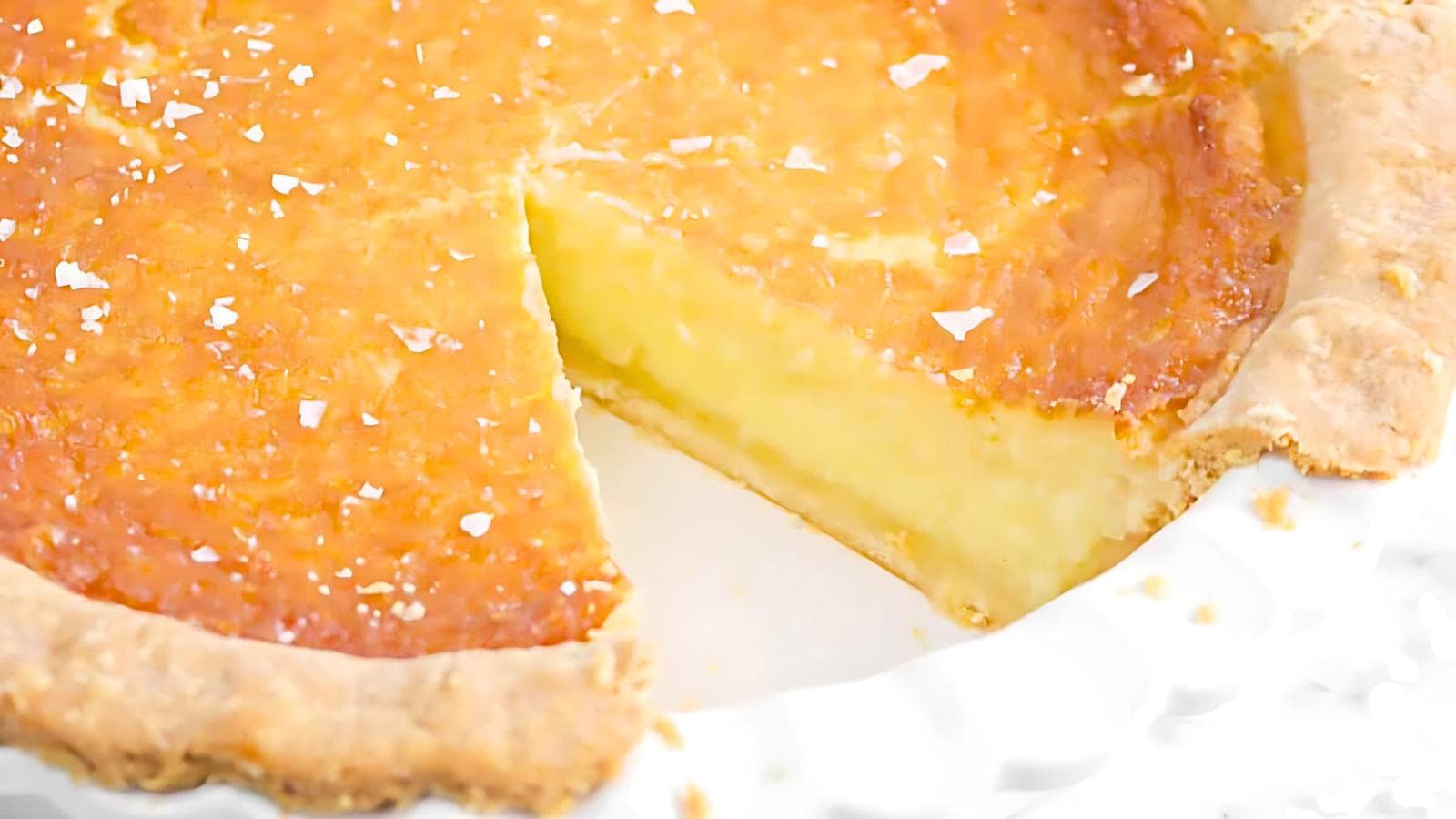 A close-up image of a creamy custard pie with a golden-brown crust. One slice has been cut and removed, revealing the smooth, pale yellow filling. The top of the pie has a slight sheen and a few sprinkled flakes of sea salt.