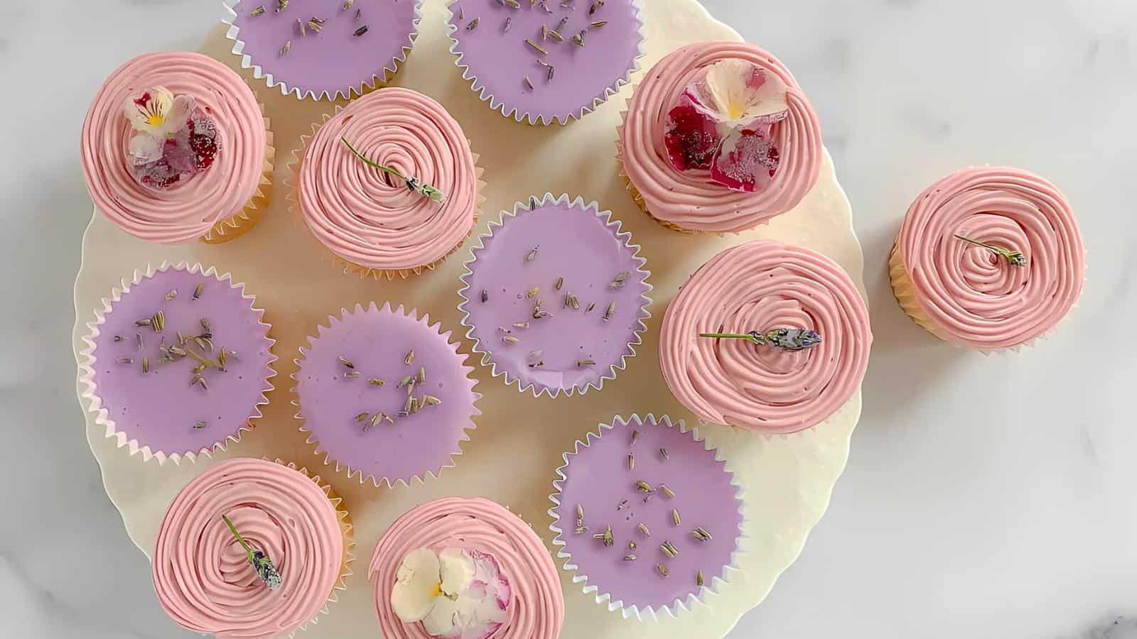 A white platter with frosted cupcakes, featuring pink swirled tops and purple flat tops, garnished with small flowers and herbs, on a light surface.