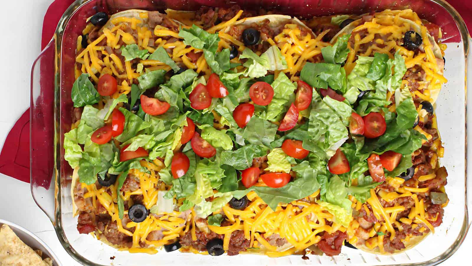 A glass baking dish with shredded cheese, lettuce, chopped tomatoes, and sliced black olives.