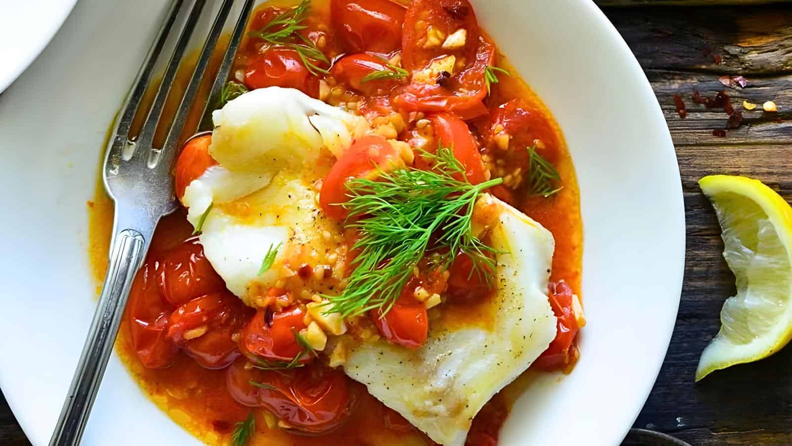 A piece of white fish topped with tomato-based sauce, garnished with fresh dill, served on a white plate with a lemon wedge and a fork.